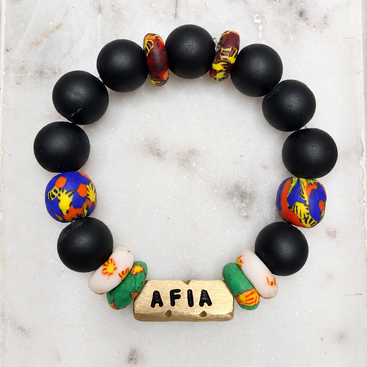 Afia Ghanaian Day Name bracelet with matte black and multicolor Krobo glass beads and an engraved brass AFIA charm on white marble.