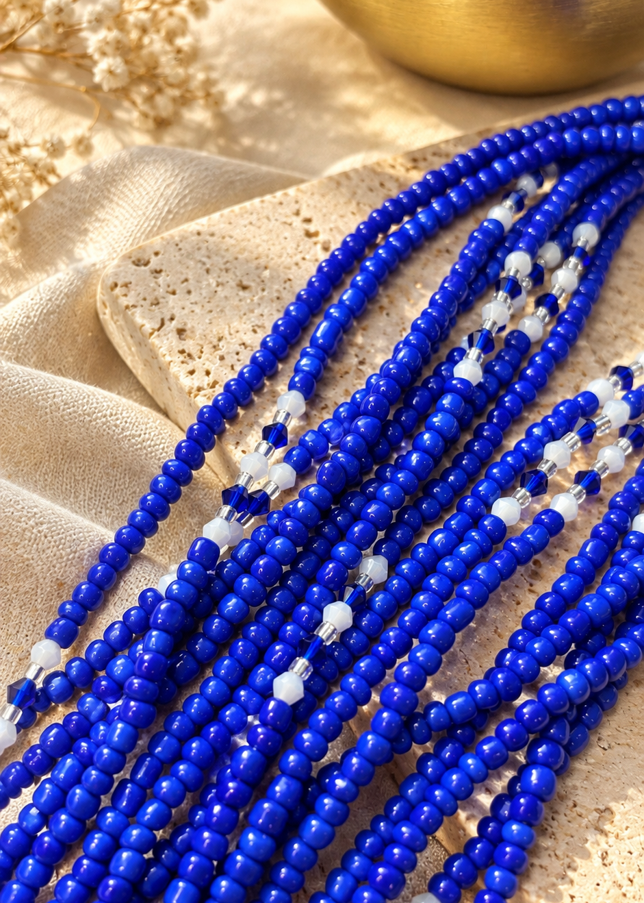 Multiple strands of royal blue faceted beads with small white accent beads, arranged on a textured beige stone and draped fabric.