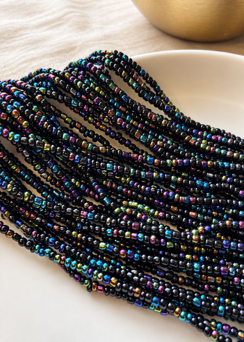 Black Girl Magic waist beads in black with iridescent jewel tones arranged on ivory fabric beside a gold vessel