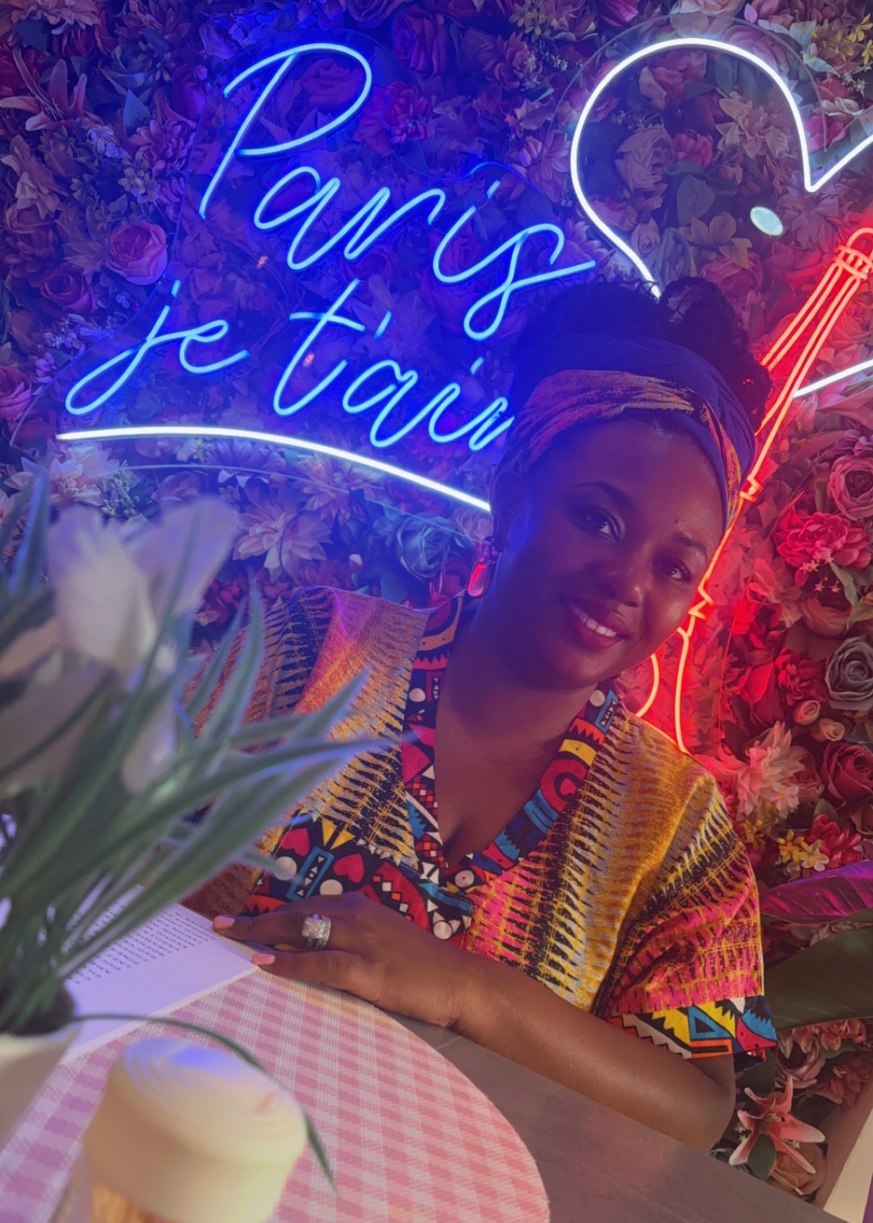 A woman wearing a colorful patterned kaftan top and headwrap sits at a table under a glowing blue neon sign that reads 'Paris je t'aime' against a floral wall.