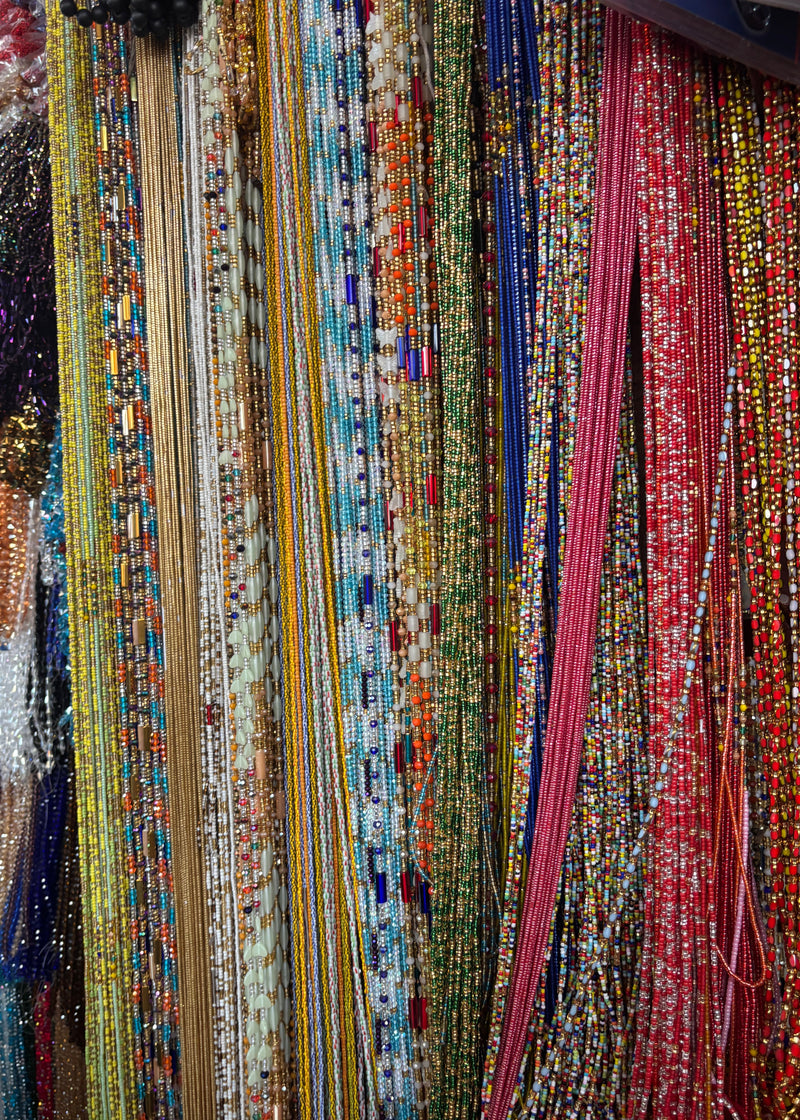 A close-up view of multiple strands of colorful waist beads hanging vertically in a display. The beads include red, pink, yellow, blue, green, white, and metallic tones, with no person or real environment visible beyond the merchandise display.