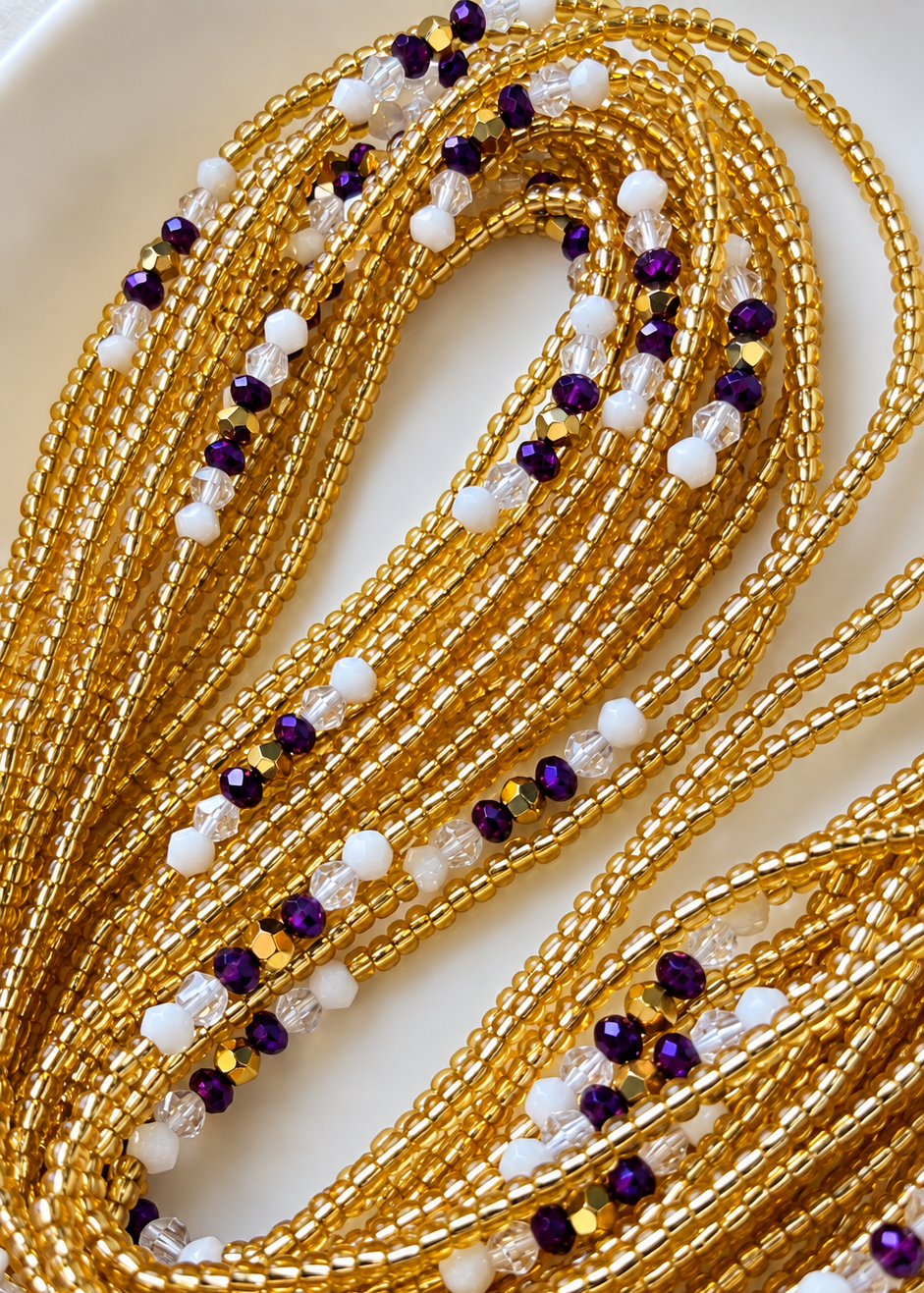 The Matriarch Luxe waist beads in gold, purple, white, and clear tones arranged on a white dish