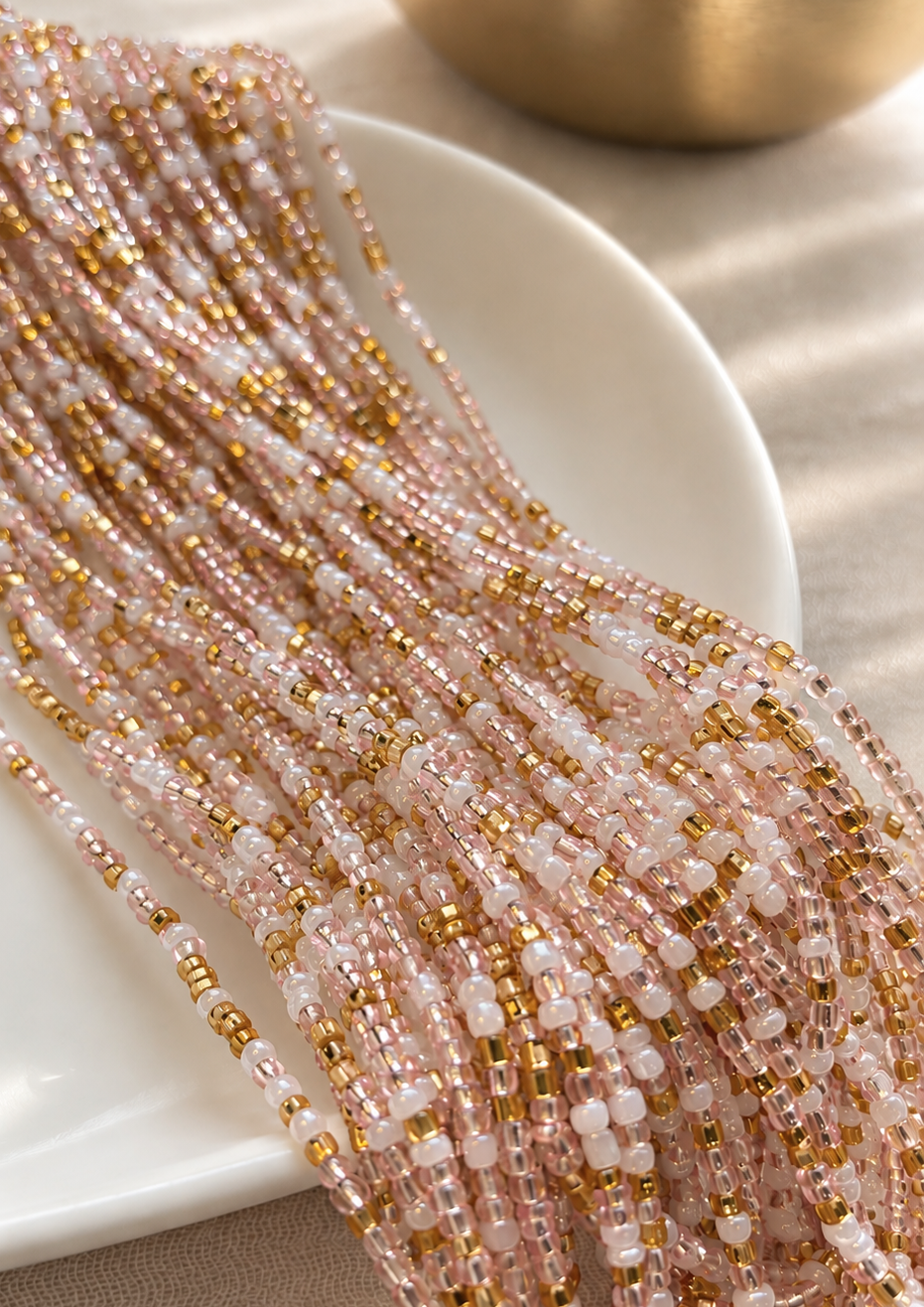 Blushing Goddess waist beads in translucent blush pink, pearl white, and gold arranged across a white dish on neutral linen