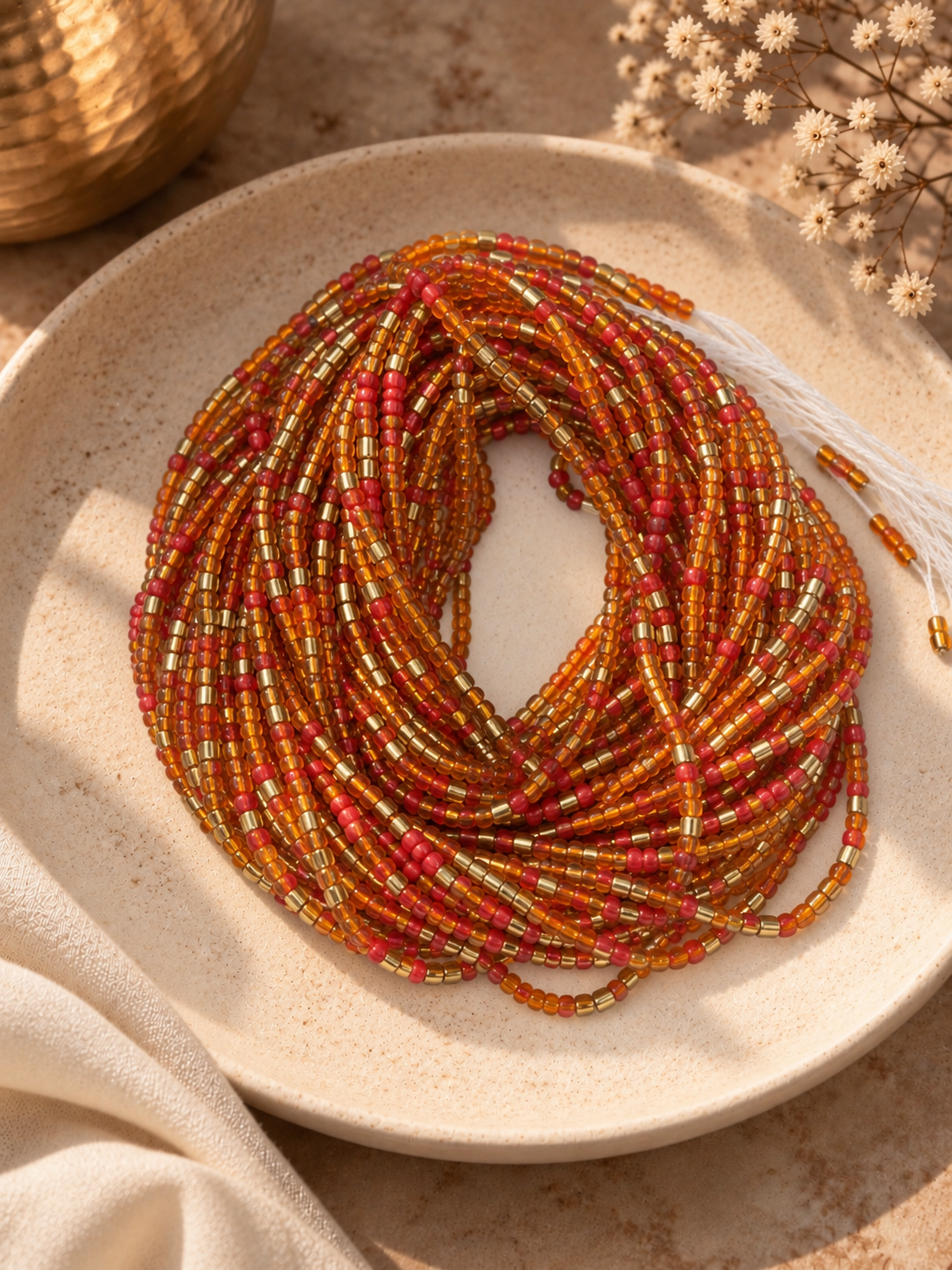Ember Sol orange, warm red, and gold waist beads arranged on a ceramic dish