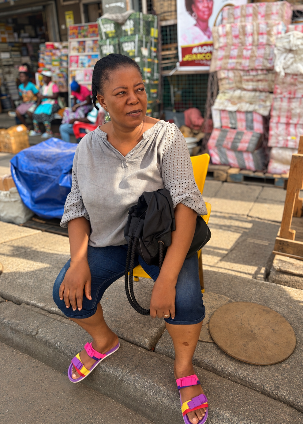 A woman sitting on a yellow chair on a street corner in front of a market stall, wearing a light gray patterned top, blue denim capris, and pink sandals, with a black shoulder bag slung over her arm.