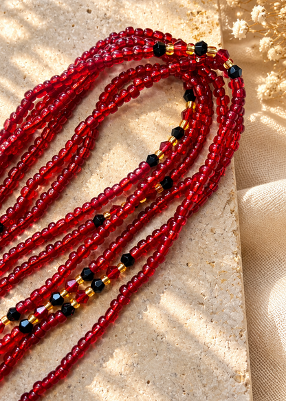 Multiple strands of red seed beads with small black and gold spacer beads, arranged on a textured beige stone slab with dried white flowers in the corner.