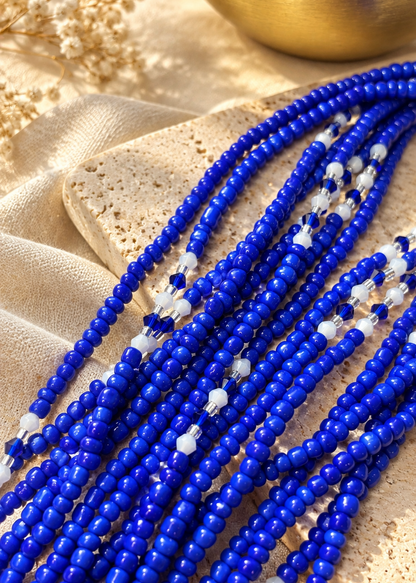 Multiple strands of royal blue faceted beads with small white accent beads, arranged on a textured beige stone and draped fabric.