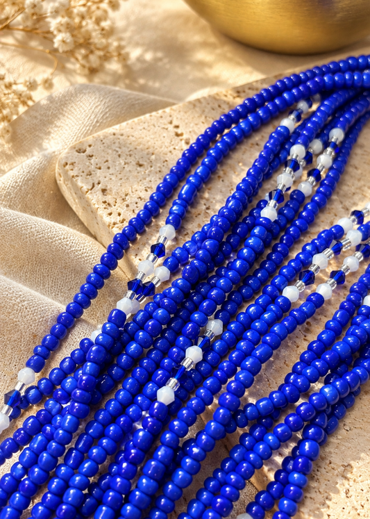 Multiple strands of royal blue faceted beads with small white accent beads, arranged on a textured beige stone and draped fabric.