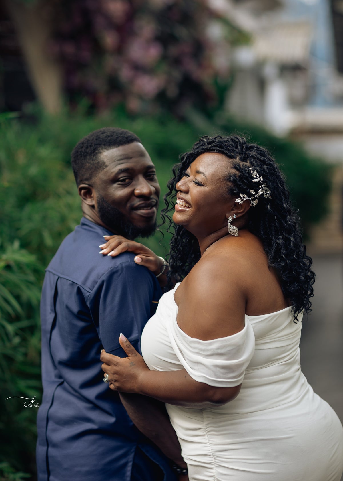 A smiling couple embracing outdoors. The woman wears an off-the-shoulder white dress and silver earrings, and the man wears a navy blue button-up shirt.